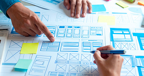 Hände arbeiten an Wireframes und Skizzen für Webseitenlayouts auf einem Tisch mit Papier, Stiften und Haftnotizen.